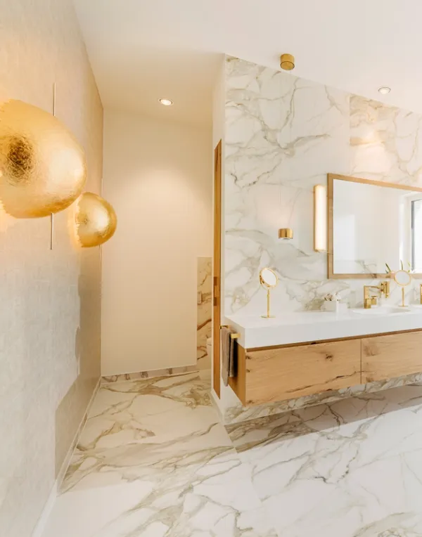 Helles Bad mit Marmor, goldener Wandleuchte und Waschtisch aus Holz vor einem großen Fenster